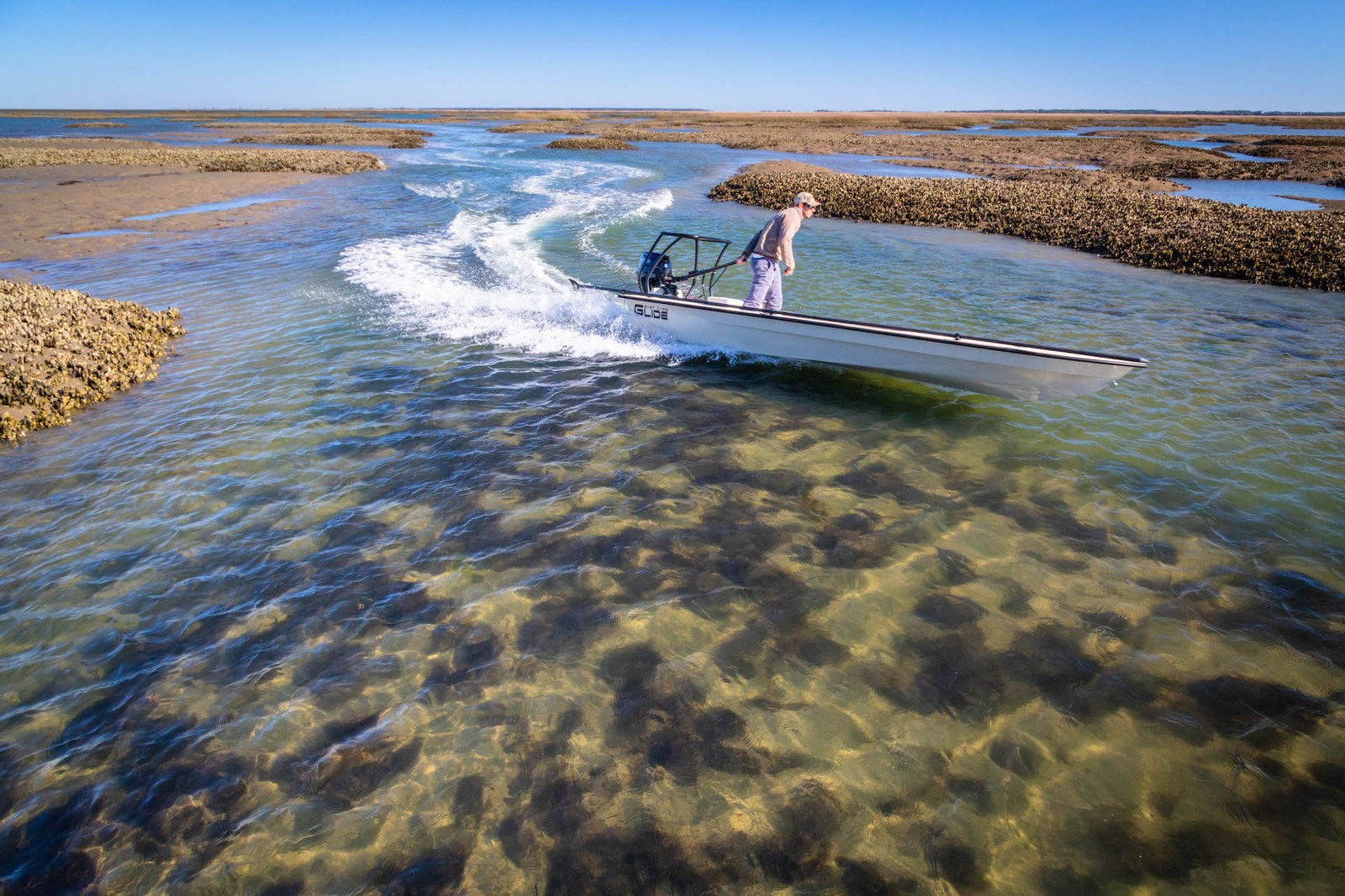 Glide running a low-tide creek between the oyster bars — tiller in hand, inches under the hull